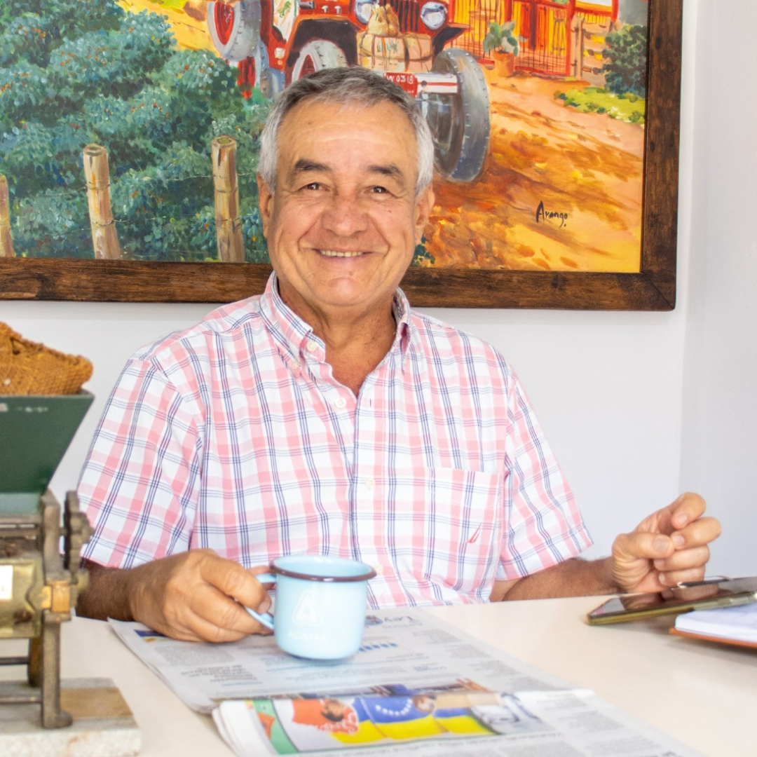 Man sitting at a table with a newspaper and mug, smiling in front of a painting.